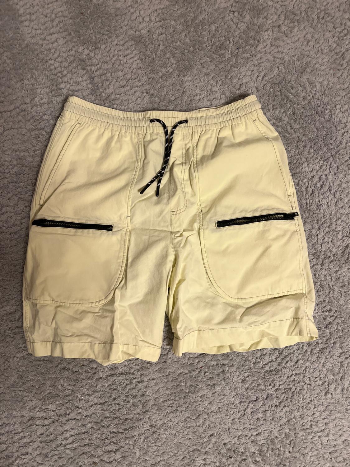 Beige shorts with black drawstring and black zip pockets, on a grey rug.
