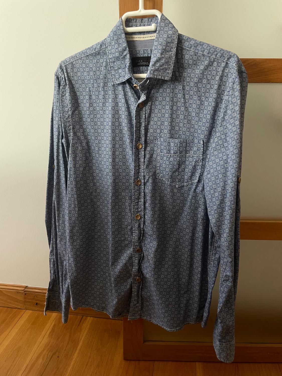 Blue long-sleeved geometric print shirt with buttons, hanging on a hanger against a wooden structure. Text: ZARA.