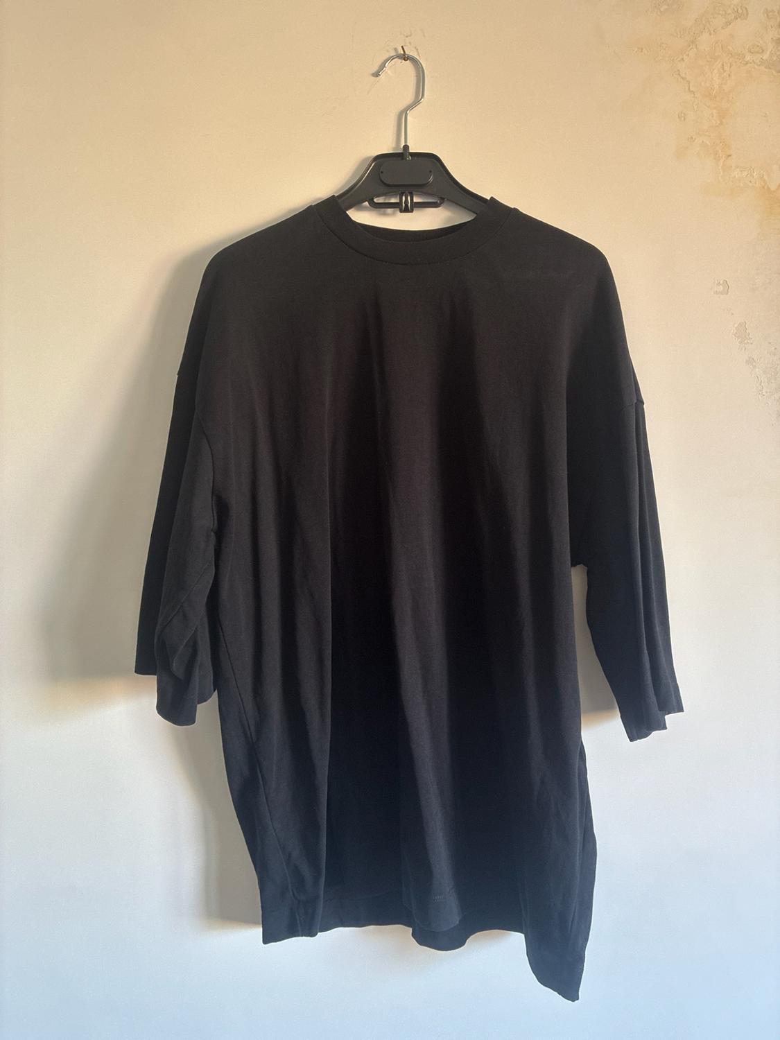 Μαύρο oversize κοντομάνικο μπλουζάκι κρεμασμένο σε μια μαύρη κρεμάστρα μπροστά από έναν ανοιχτόχρωμο τοίχο με υφή. Casual και χαλαρό στυλ.