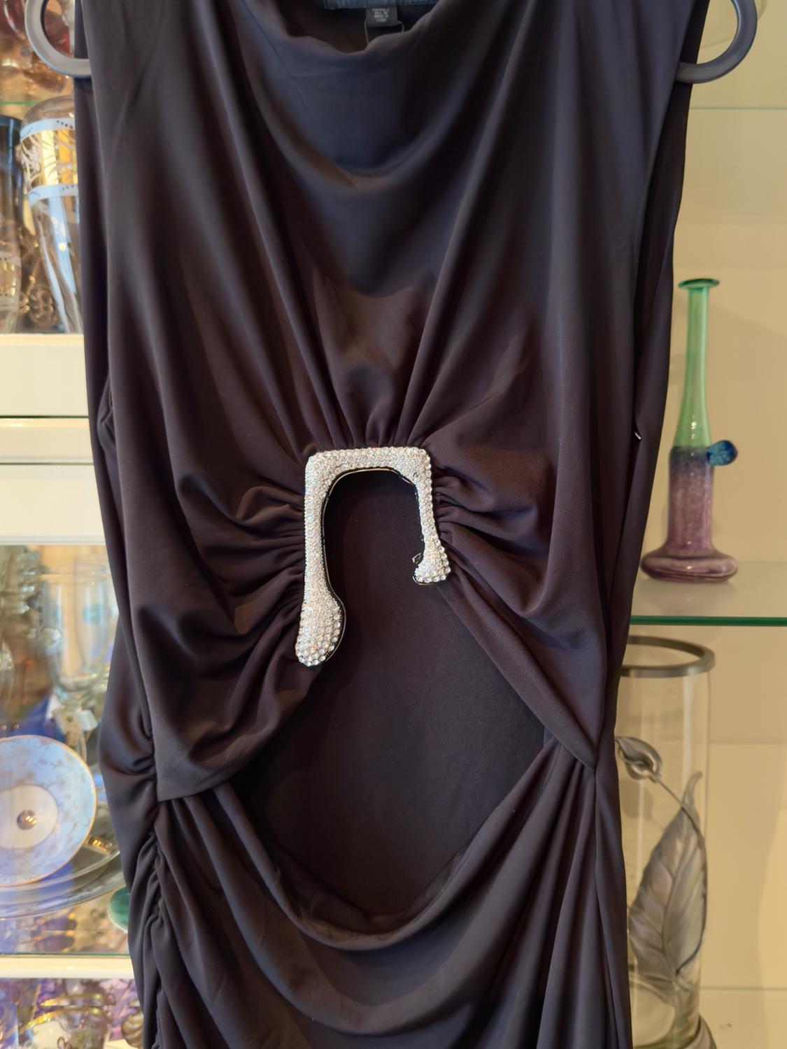 Black draped dress on a hanger with a shiny silver embellishment in the centre, in front of a glass display cabinet with decorations.