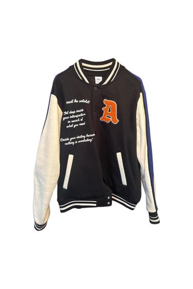 Black bomber jacket with white sleeves, an orange letter A patch, embroidered “must be untold” text and striped details.