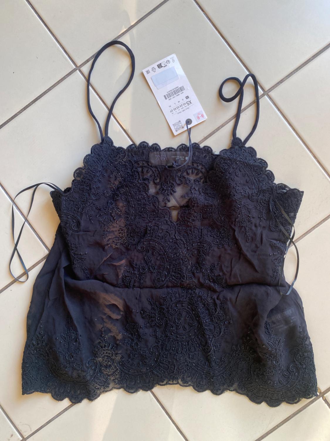 Semi-sheer black top with embroidery and spaghetti straps on light tiles. Label with text ZARA XS.