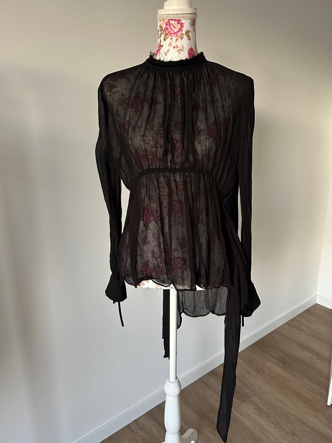 Semi-sheer black blouse with floral print on a mannequin with a white base and decorated neckline, on a wooden floor.