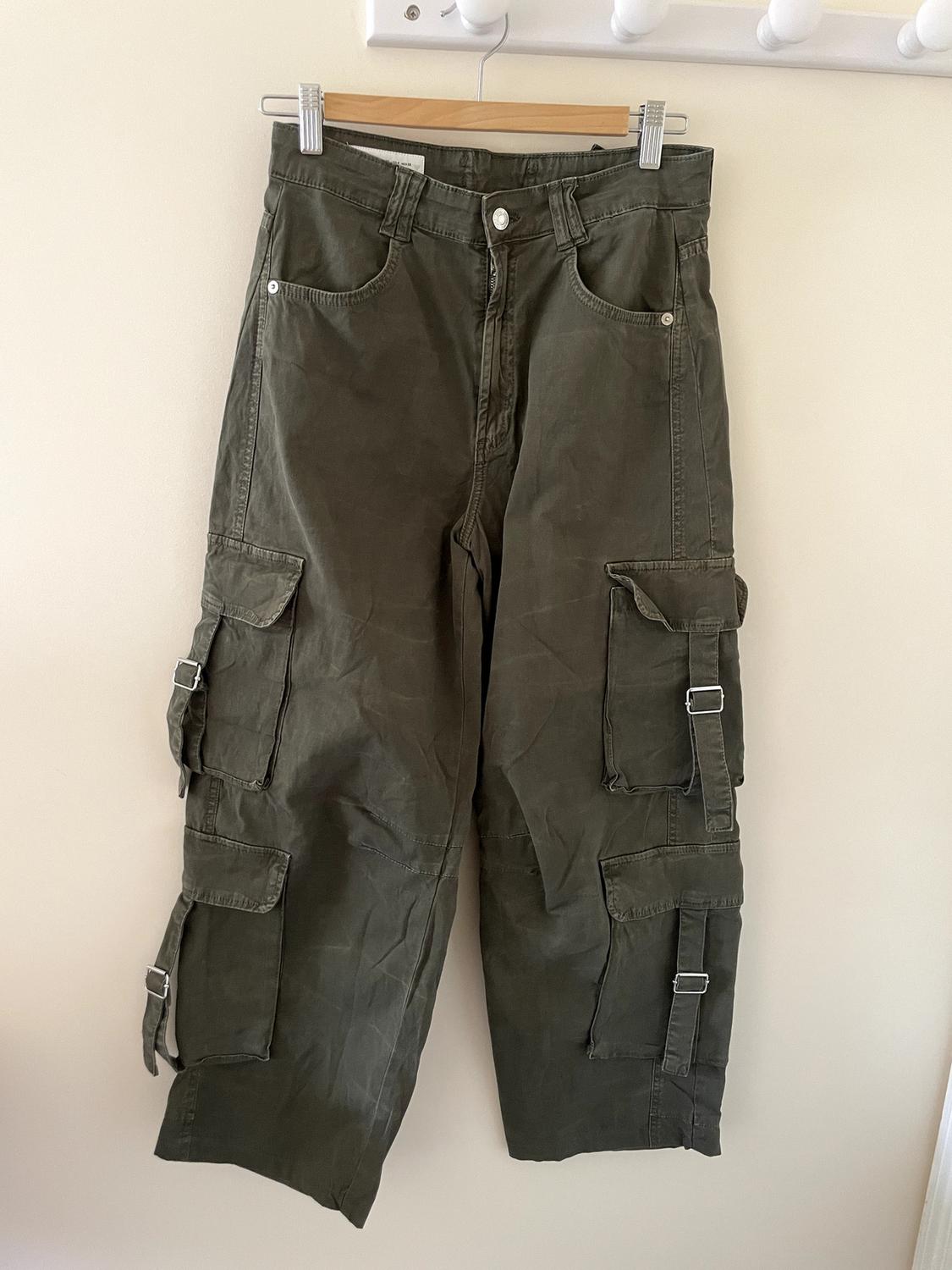 Olive green cargo trousers hanging on a wooden hanger against a light background. Their side pockets with straps and buckles stand out.