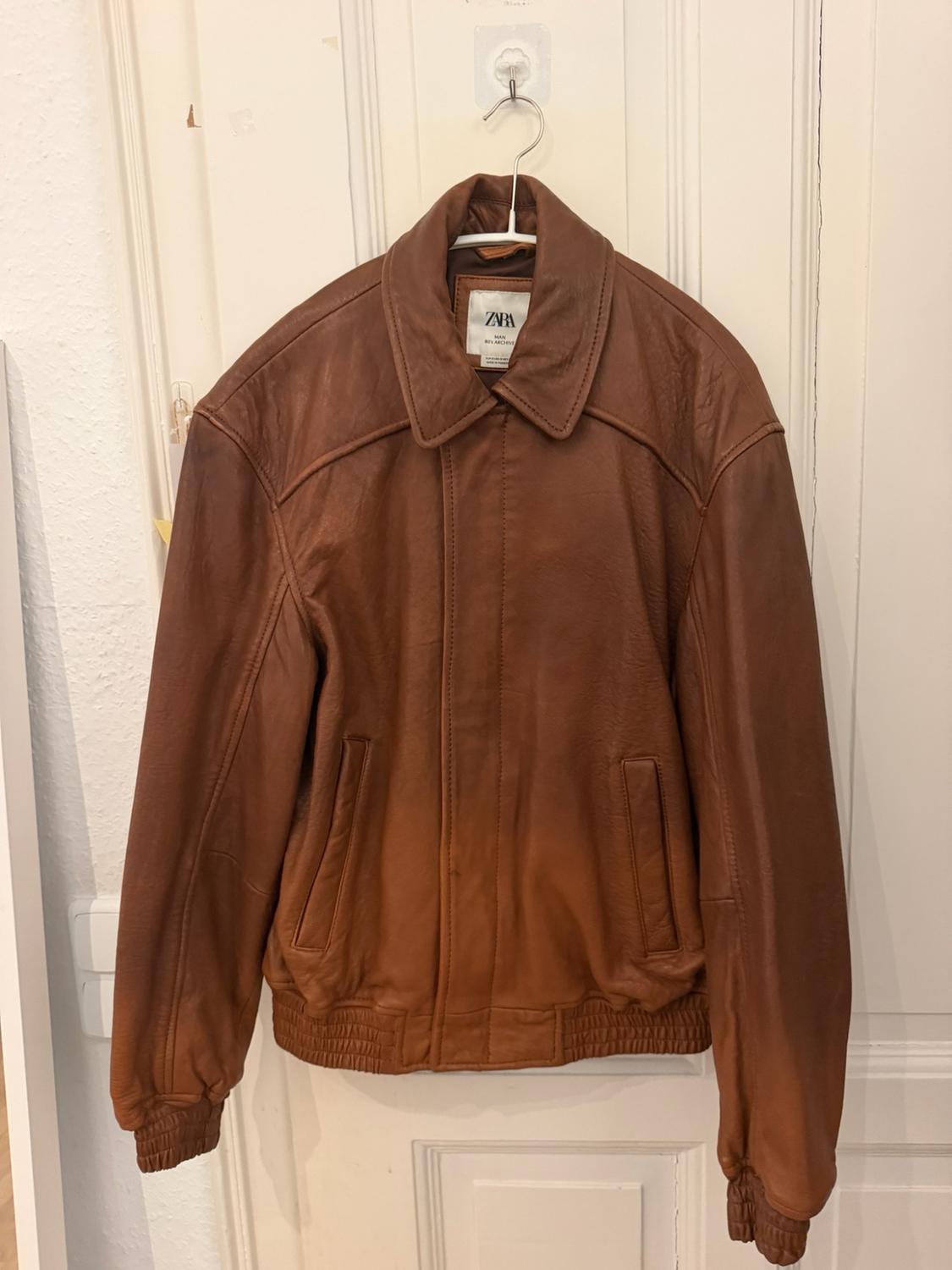 Brown leather jacket hanging on a white door. It has a collar, pockets and elasticated cuffs. Text: ZARA MAN.