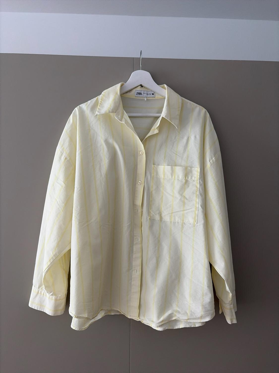 ZARA loose-fit long-sleeved shirt with fine yellow stripes and a pocket, hanging on a white hanger against a neutral grey background.