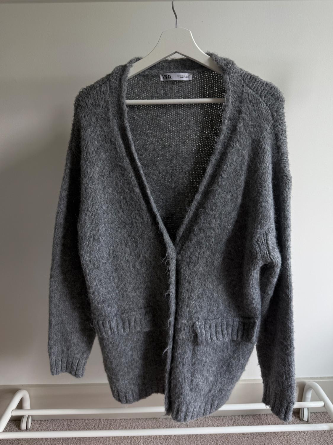 Chunky knit grey cardigan with front pockets, hanging on a white hanger against a neutral backdrop. Visible label: Zara.