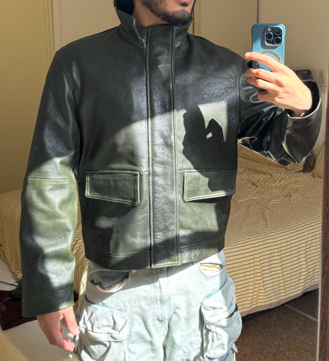 Mirror selfie of a man in his bedroom. He is wearing a dark green leather jacket and light denim cargo trousers.