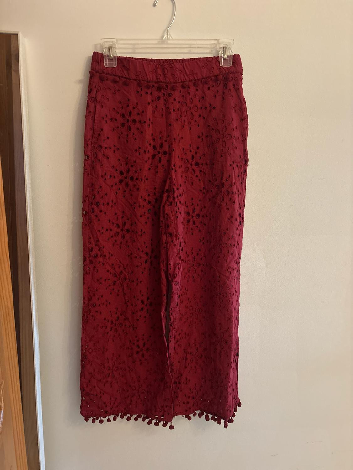 Maroon wide-leg pants with a floral openwork design and pom-poms at the waist
and hem, hanging on a clear hanger against a white wall.
