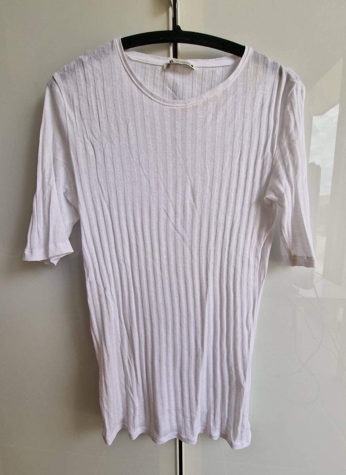 Βασικό λευκό ριμπ t-shirt με κοντά μανίκια, κρεμασμένο σε μαύρη κρεμάστρα σε ανοιχτόχρωμο φόντο.