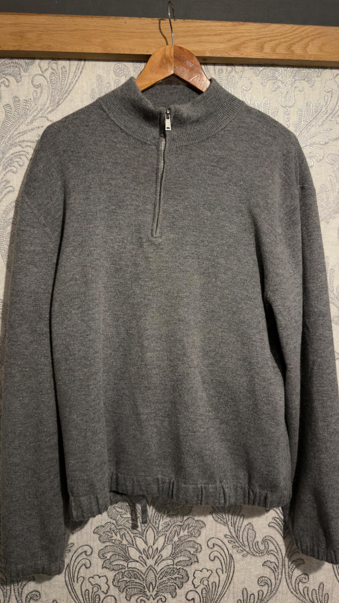Grey knitted half-zip jumper with an elasticated hem, hanging on a wooden hanger against a floral wallpaper background.