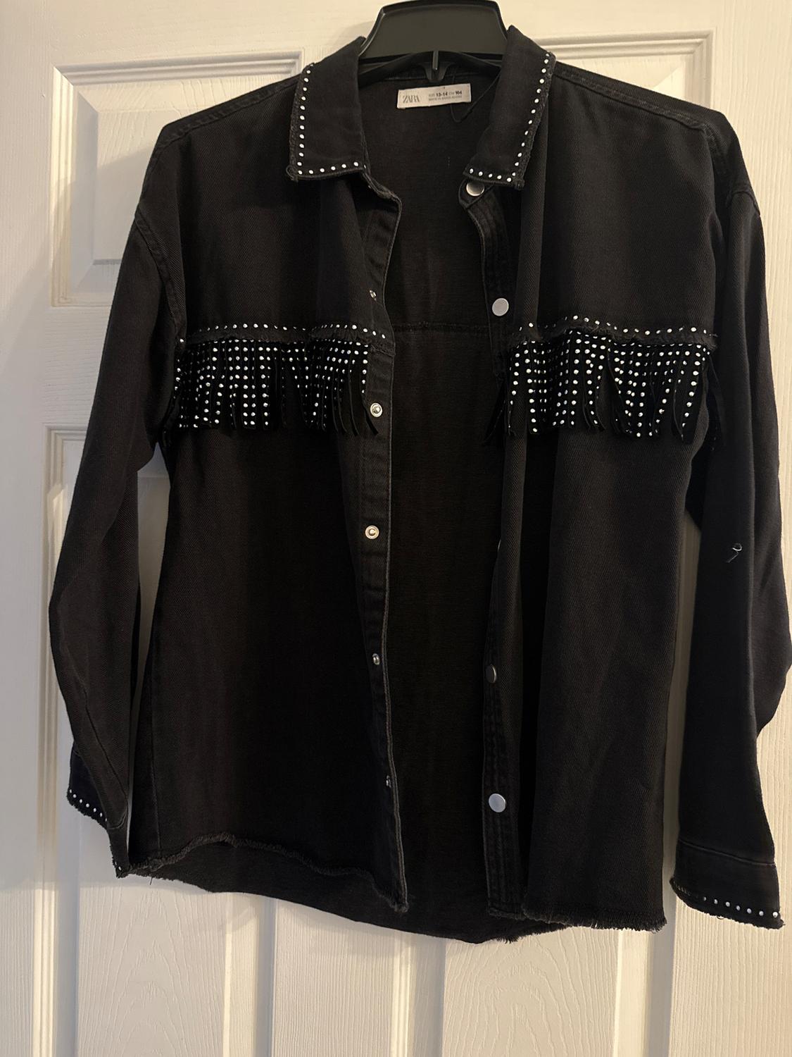 Black shirt with fringes and studs on the chest and collar, hanging on a white door. Visible tag on the collar.
