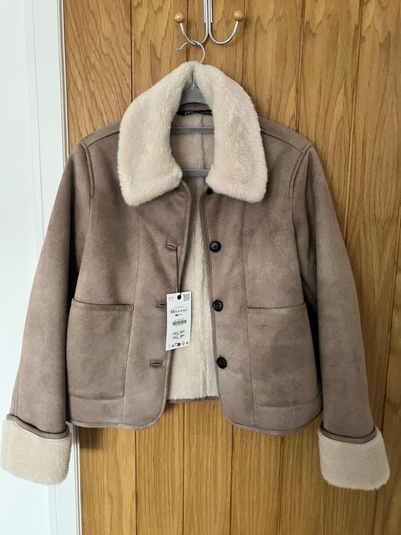 Brown jacket with white fleece lining and collar, hanging on a wooden hanger. Label visible on the front.