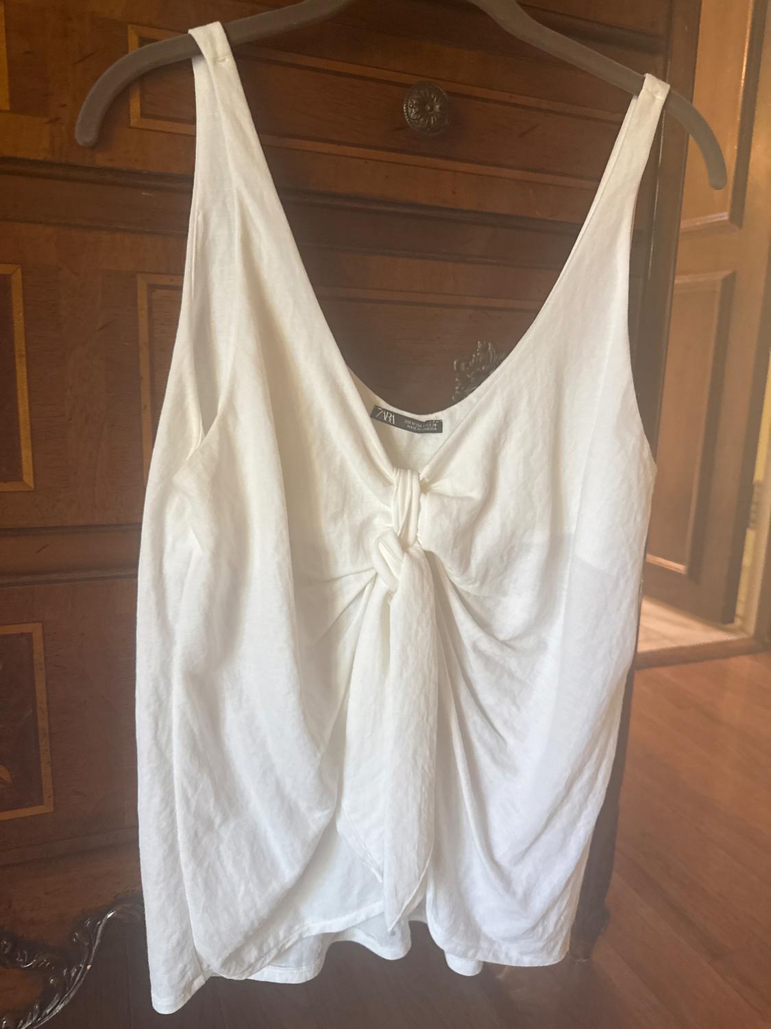 White sleeveless blouse with front knot hanging on a hanger, wooden furniture background. Visible tag: ZARA.