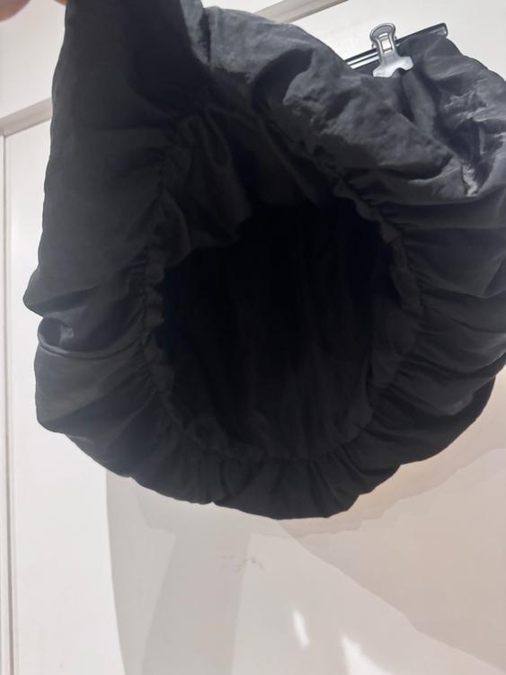 Black bag with a ruched design hanging from a hanger against a white background. The light highlights the material's textures.