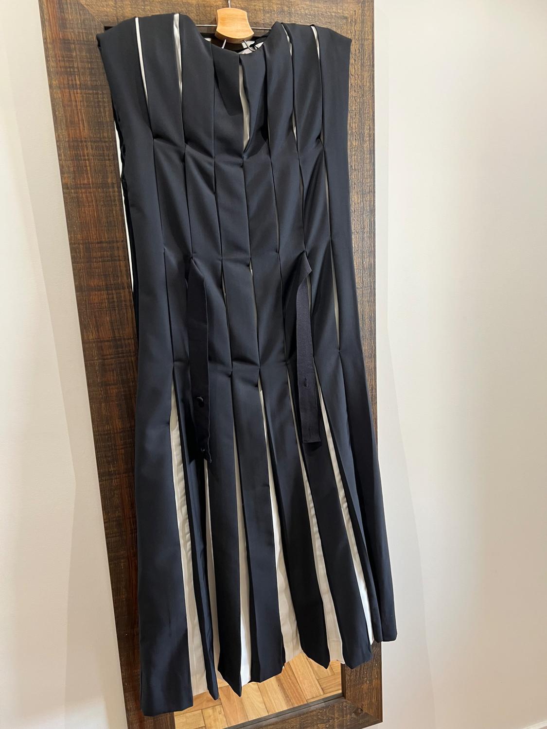 Black dress with vertical pleats and white details, hanging on a wooden clothes rail against a light background.