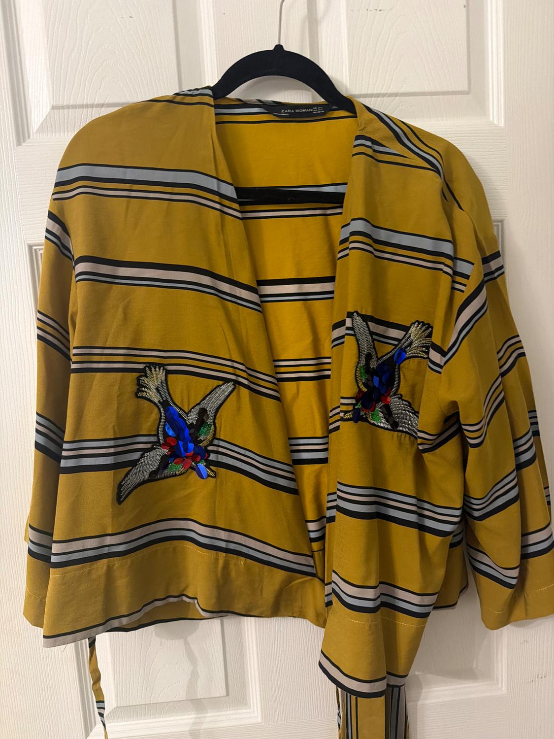 Yellow jacket with black and white stripes, bird embroidery on the chest. Hanging on a black hanger in front of a white door.