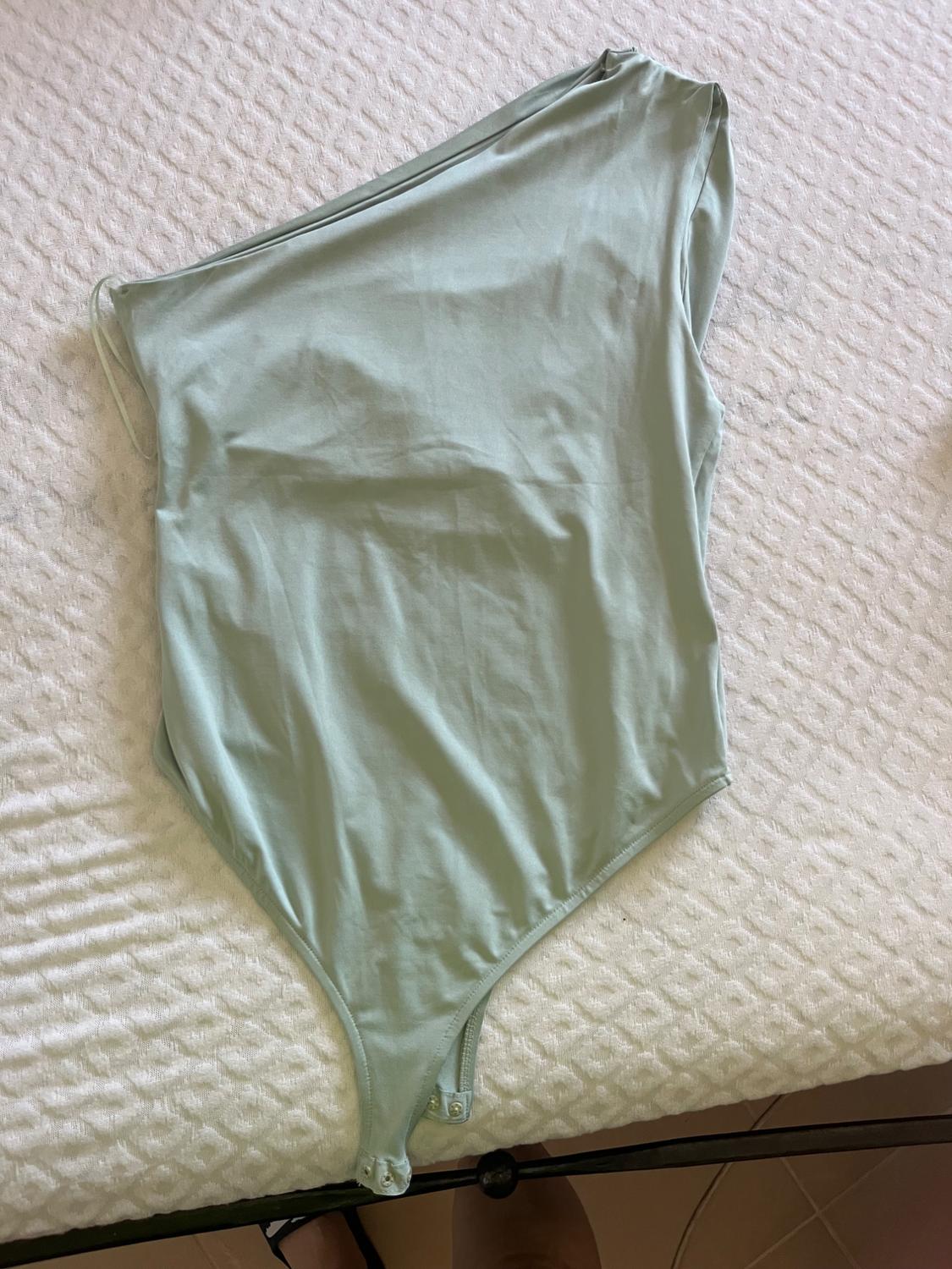 Light green asymmetrical one-shoulder bodysuit, laid out on a textured white surface in a relaxed style.