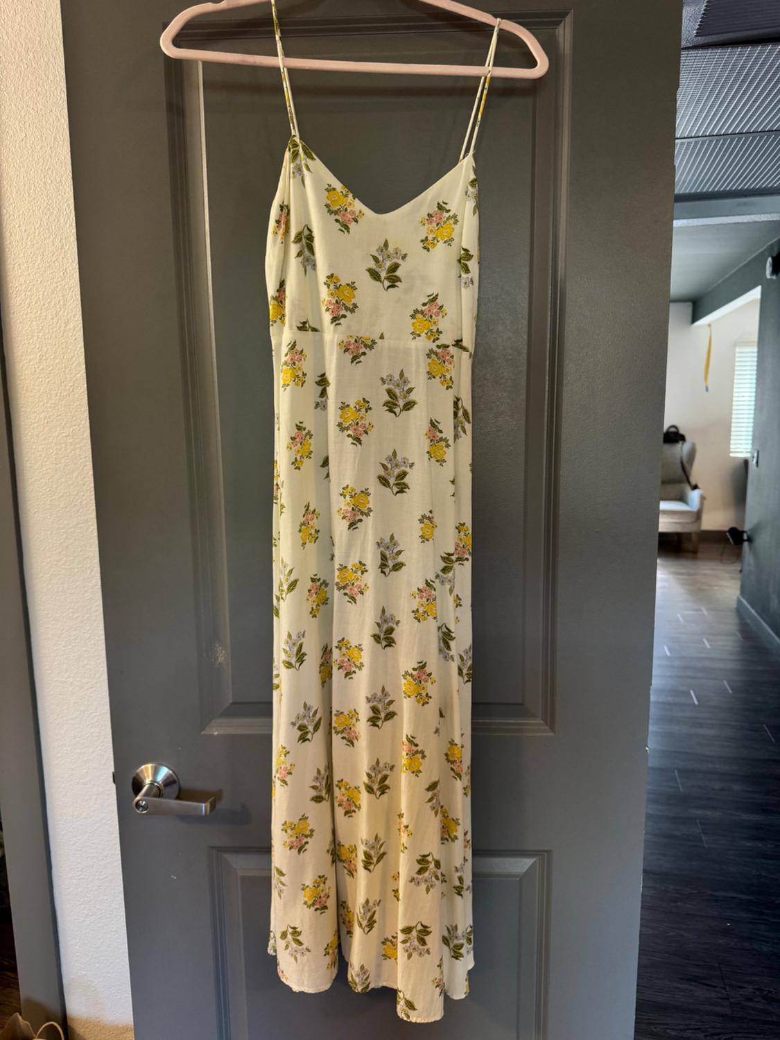 A long, spaghetti-strap dress with a yellow and green floral print, hanging on a coat hanger over a grey door.