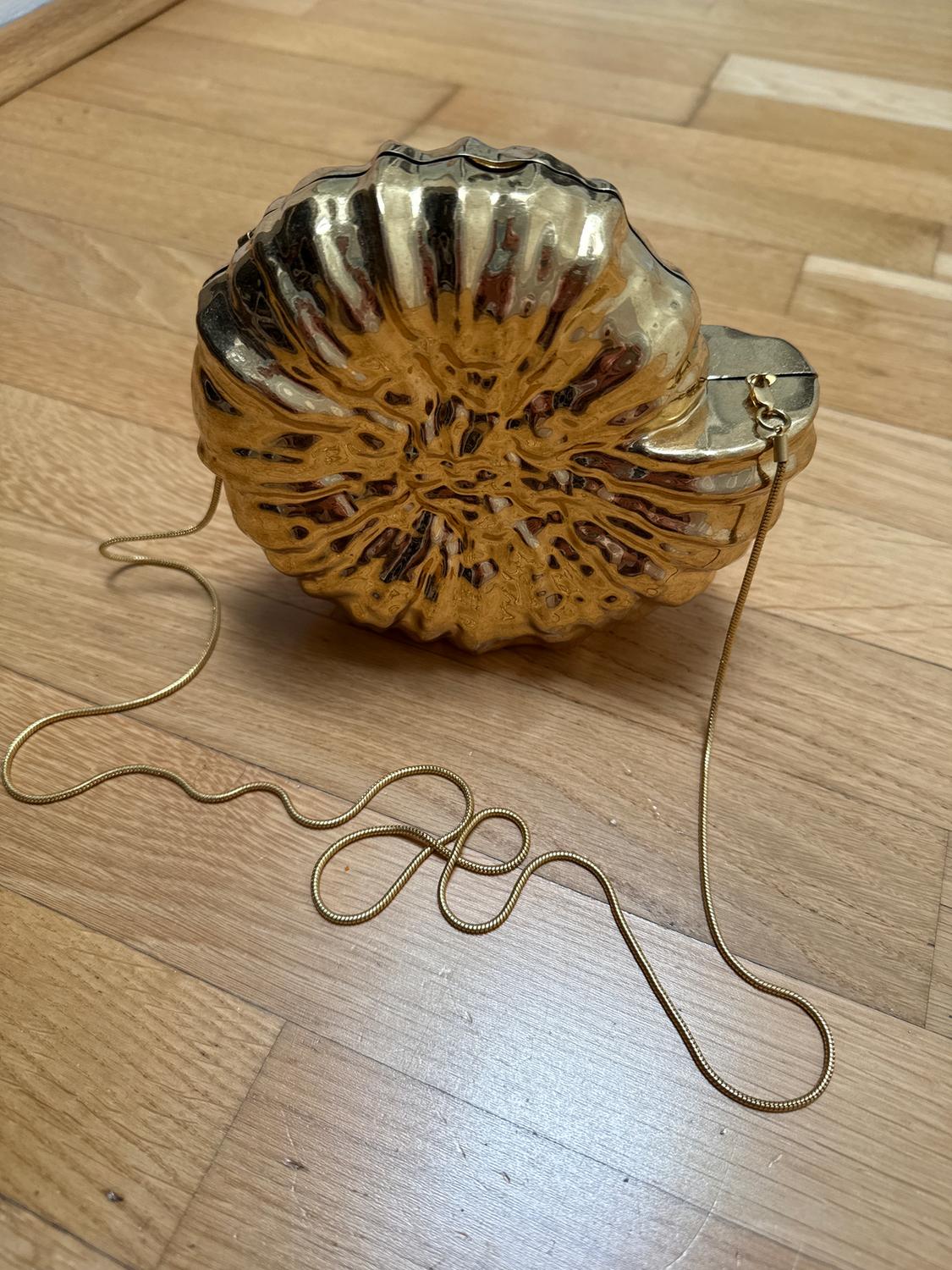 Gold shell-shaped handbag on a wooden floor. Gold chain extended in a spiral around the handbag.