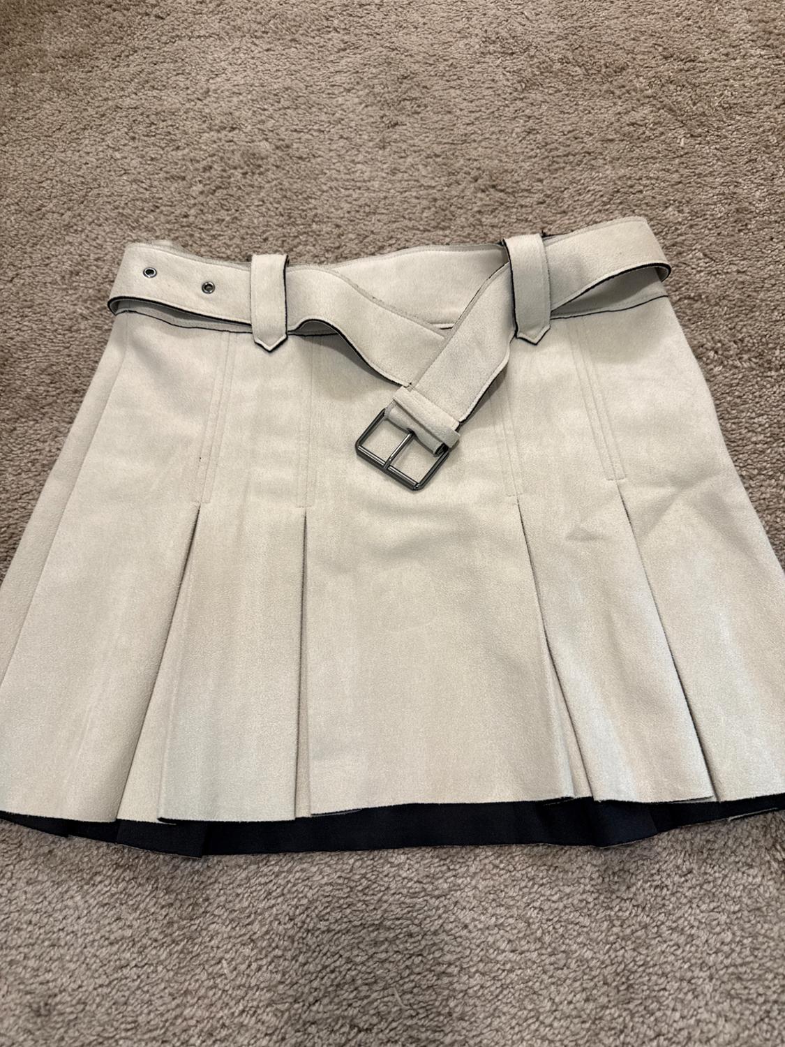 Beige pleated miniskirt with belt and buckle, black interior visible at the hem, laid out on a rug.