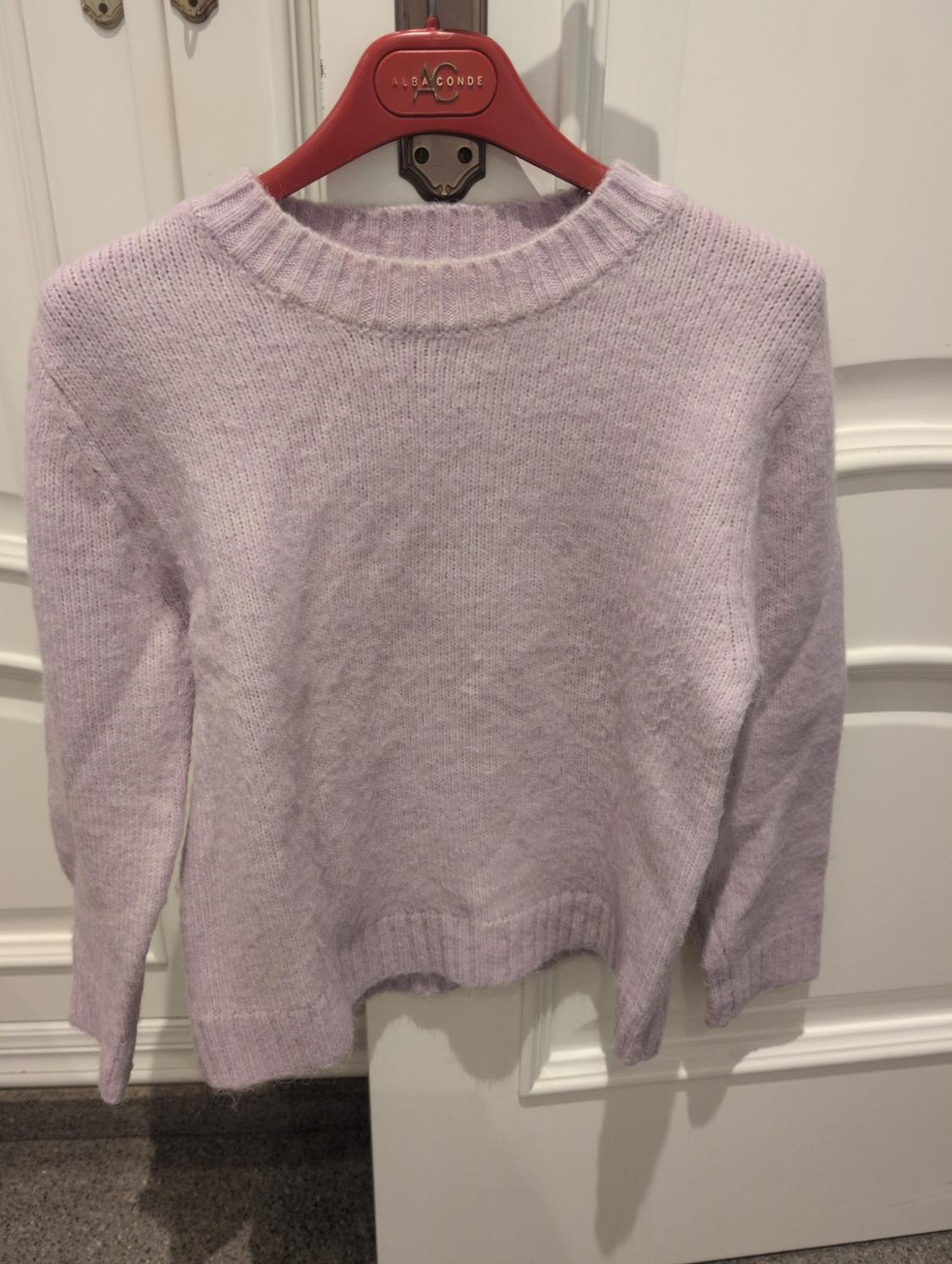 Lilac knitted jumper hanging on a red "ALBA CONDE" branded hanger, against a background of white doors.