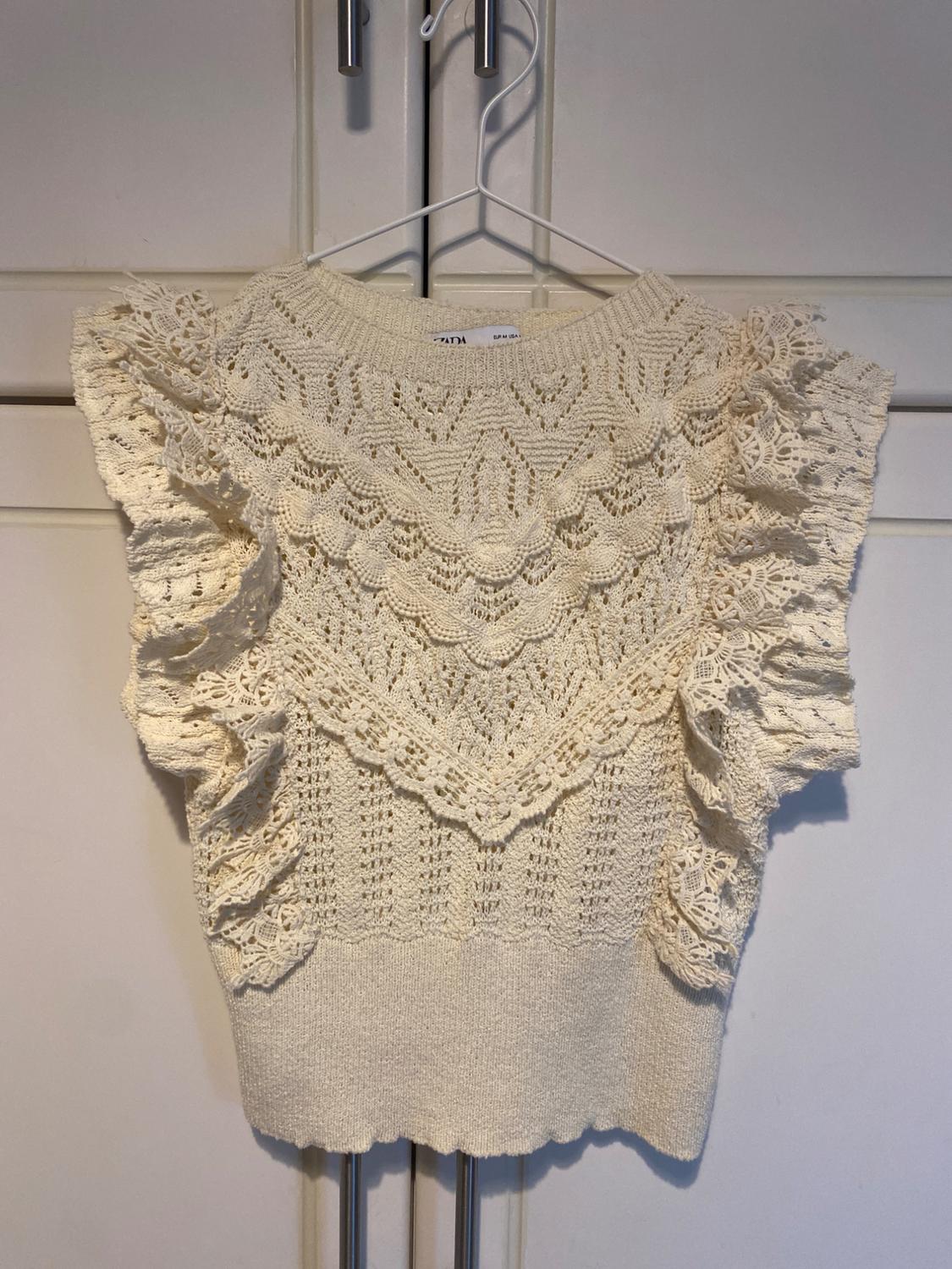Ecru open-knit top with ruffles, hanging on a white hanger in front of a wardrobe. Label with ZARA text.