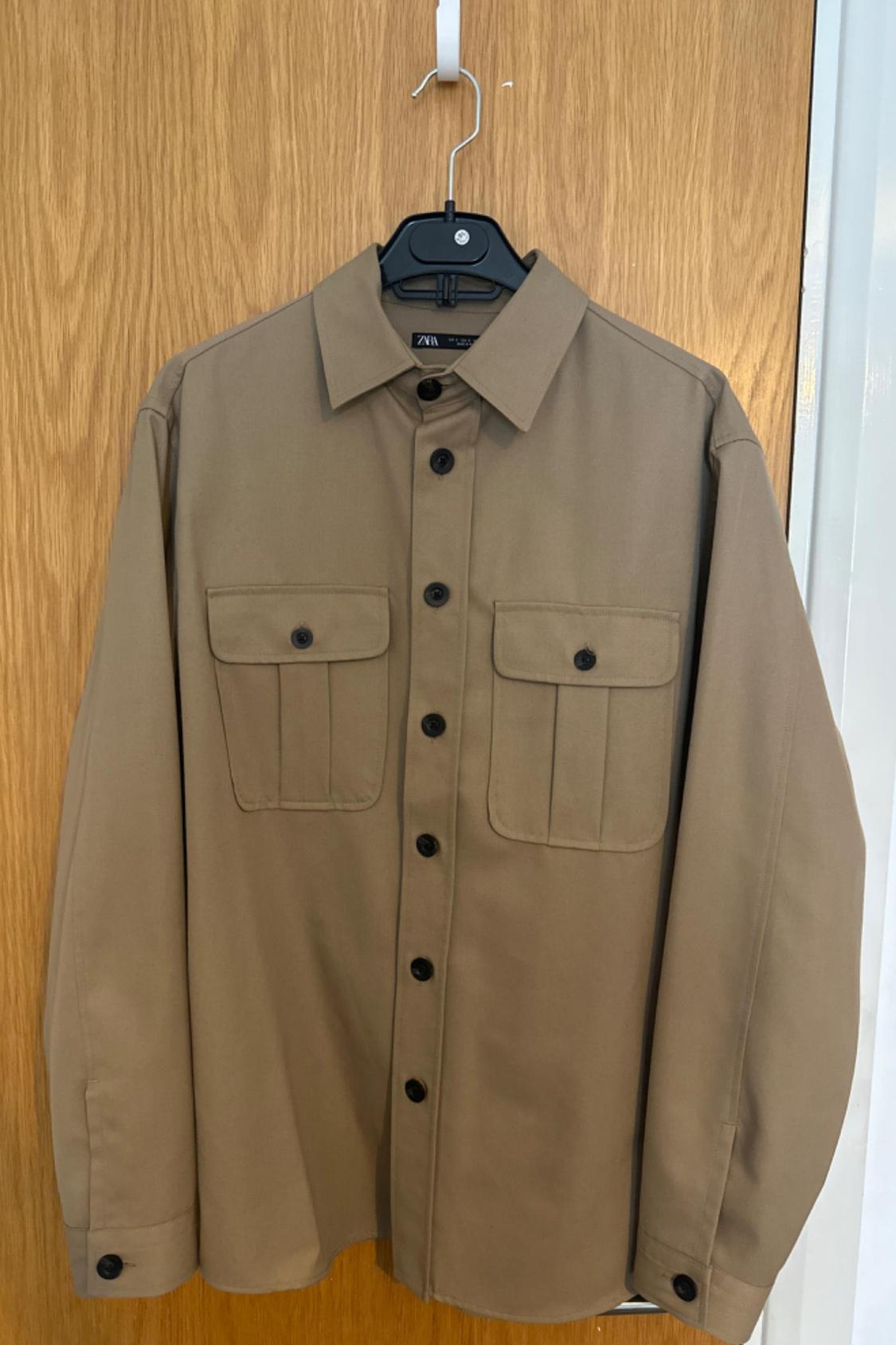 Front view of a beige shirt with flapped front pockets and black buttons.