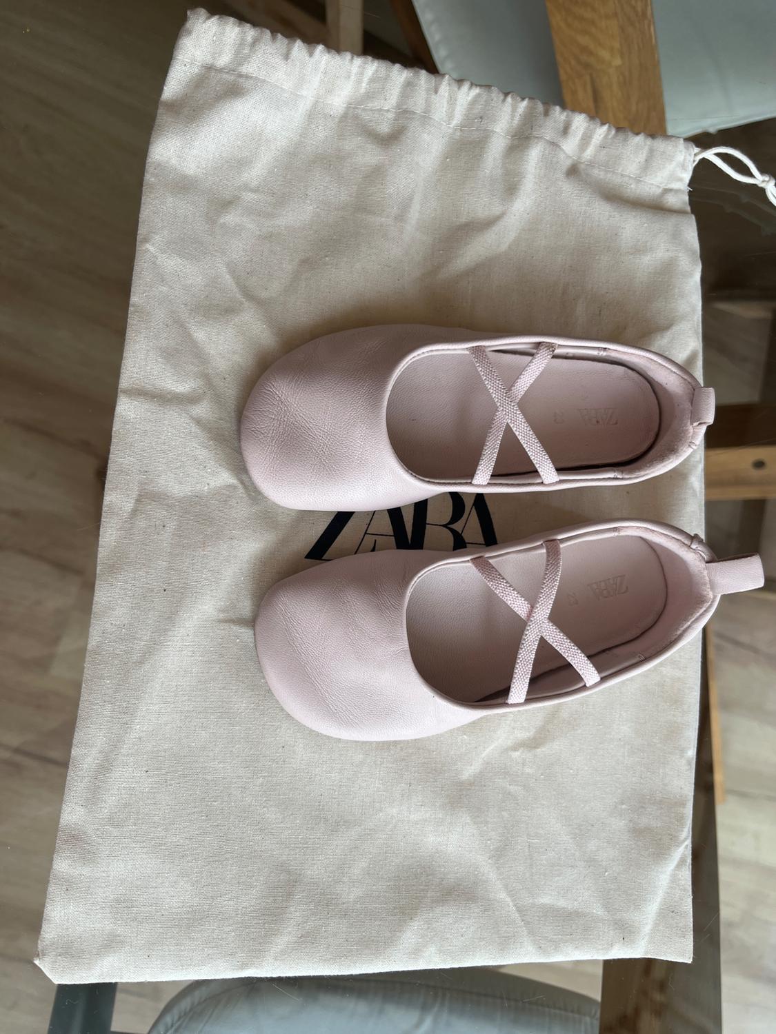 Light pink flat shoes with crossed straps on a beige fabric bag with the "ZARA" logo partially visible.