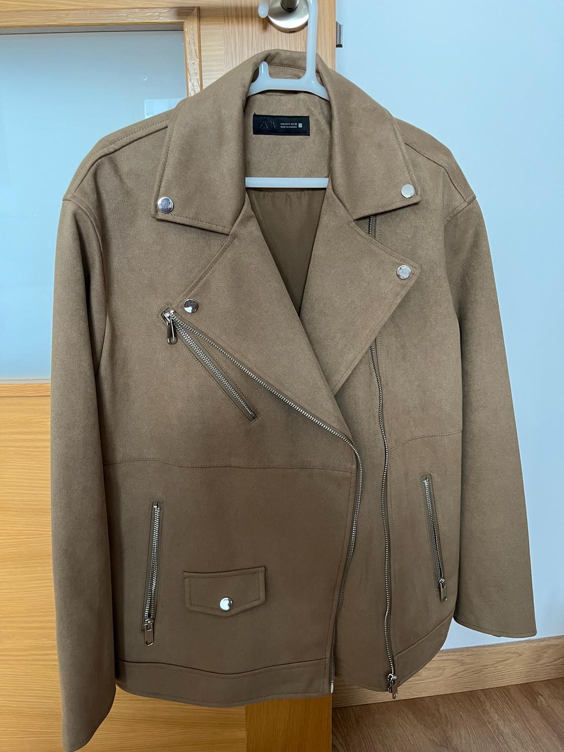 Καφέ biker jacket με ασημί φερμουάρ κρεμασμένο σε κρεμάστρα πάνω σε ξύλινη πόρτα. Ετικέτα με το κείμενο ZARA.