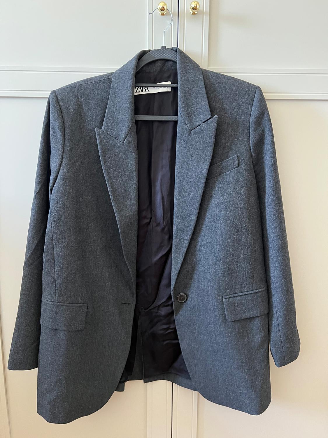 Grey blazer on a hanger in front of a white wardrobe with gold knobs. Open design with lapels, dark lining and a label with the text ZARA.