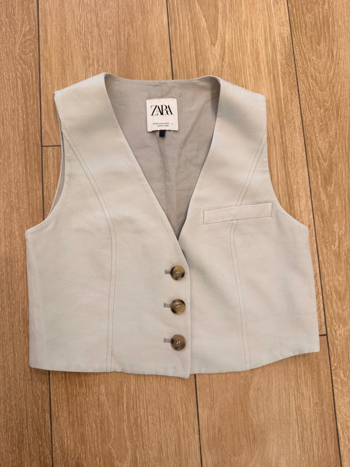 Beige V-neck waistcoat with three buttons and a chest pocket, on a wooden floor. Label with the text ZARA.