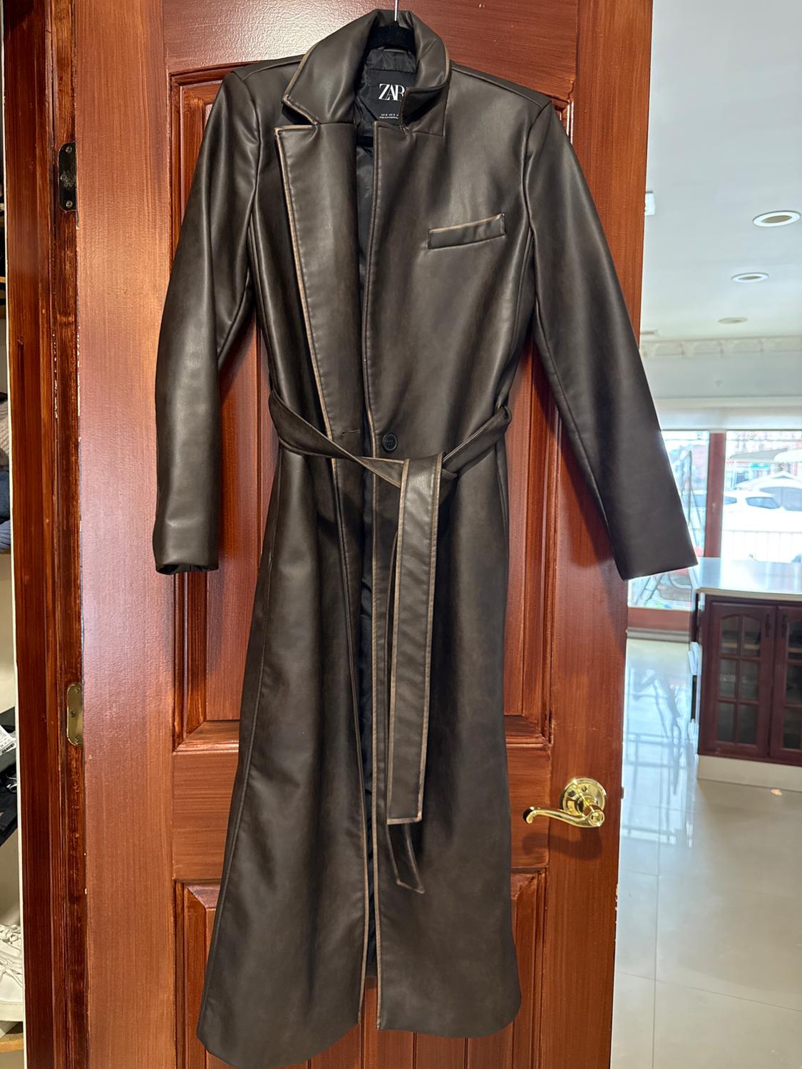 Long black coat hanging on a wooden door. It has wide lapels, a belt, and a chest pocket. "ZNR" label visible.