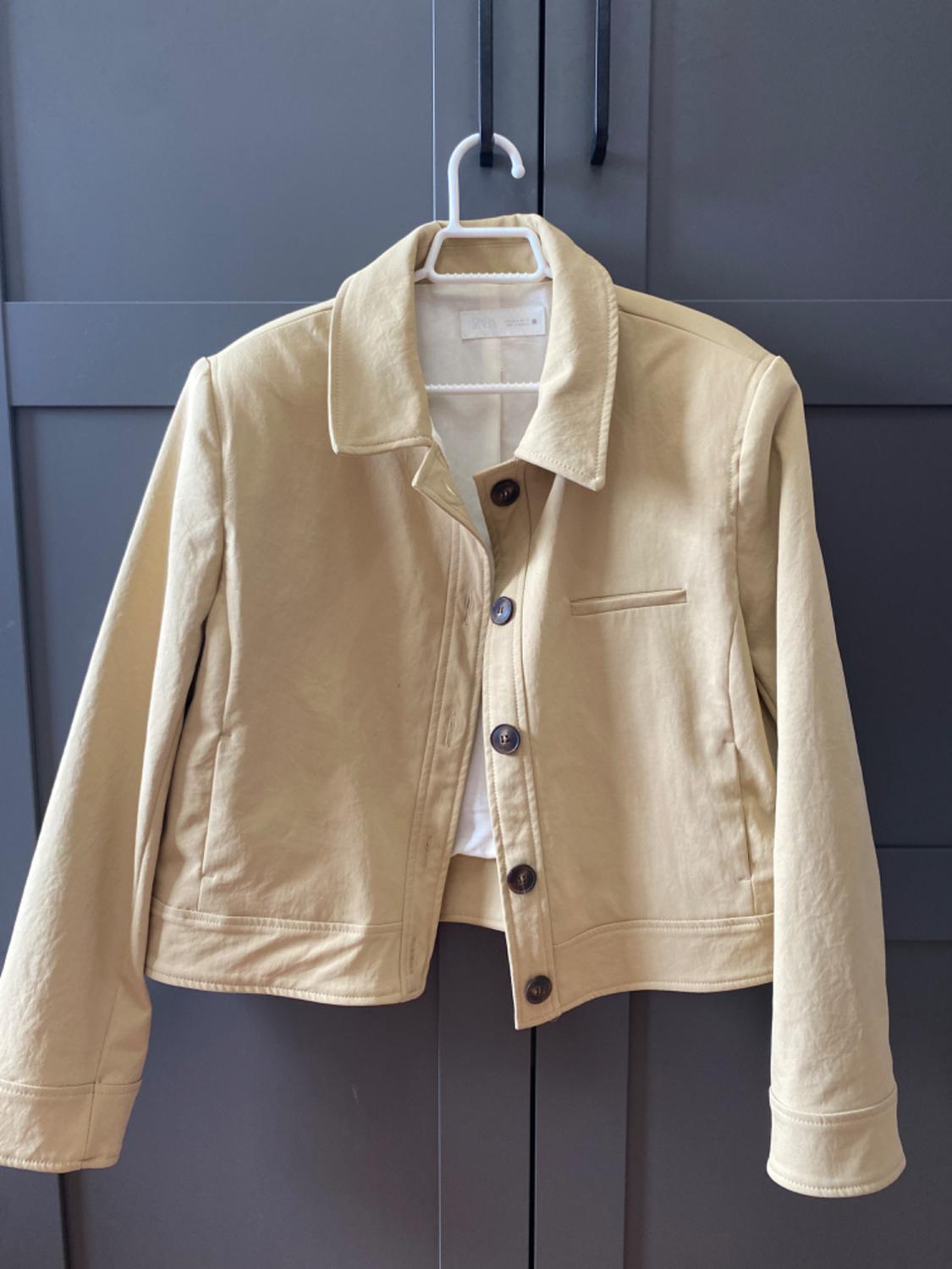 Beige jacket with dark buttons hanging on a white hanger in front of a dark grey wardrobe.