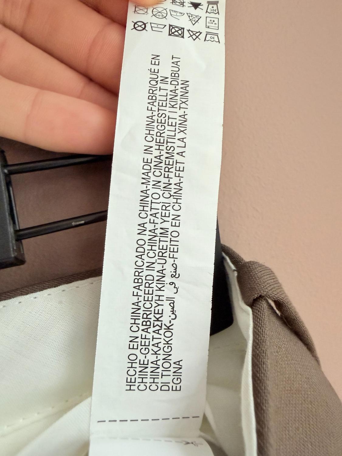 Close-up of a hand holding a label with washing symbols and the text MADE IN CHINA-FABRICADO NA CHINA-MADE IN CHINA on a brown garment.