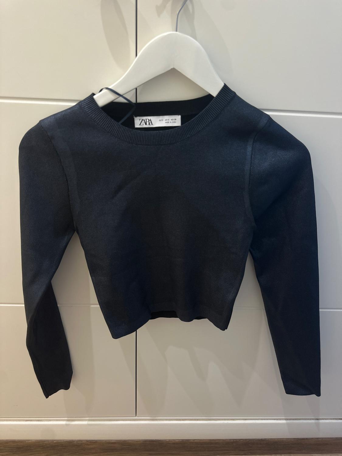 Long-sleeved black jumper hanging on a white hanger, with a visible Zara label, against a background of light-coloured tiles.