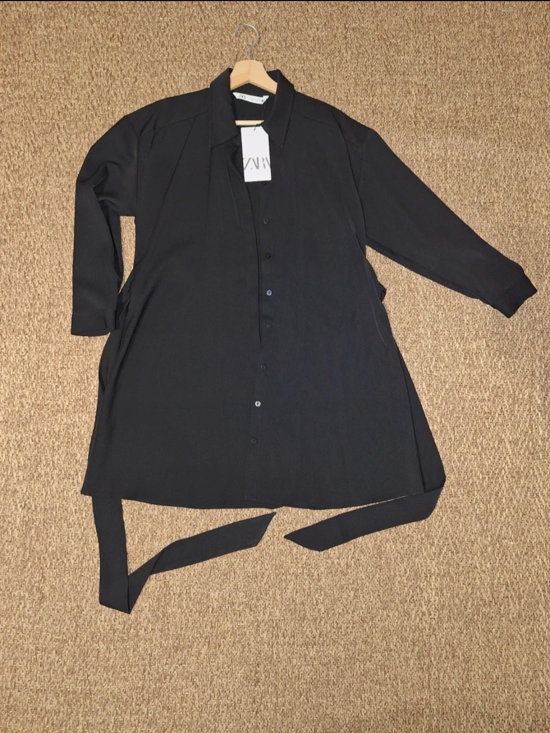 Black long-sleeved shirt with buttons and belt, hanging on a hanger over a beige carpet. Visible label with "ZARA" text.