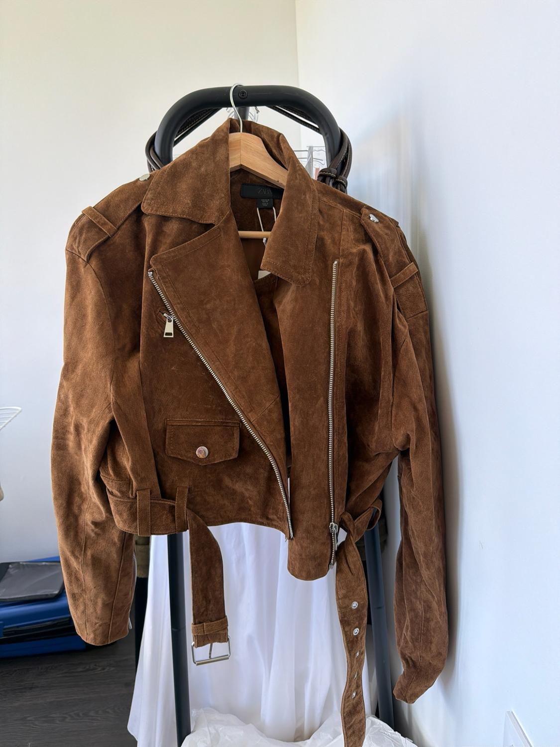 Brown biker jacket with zips and a belt, hanging on a wooden hanger above a white garment in a bright interior.