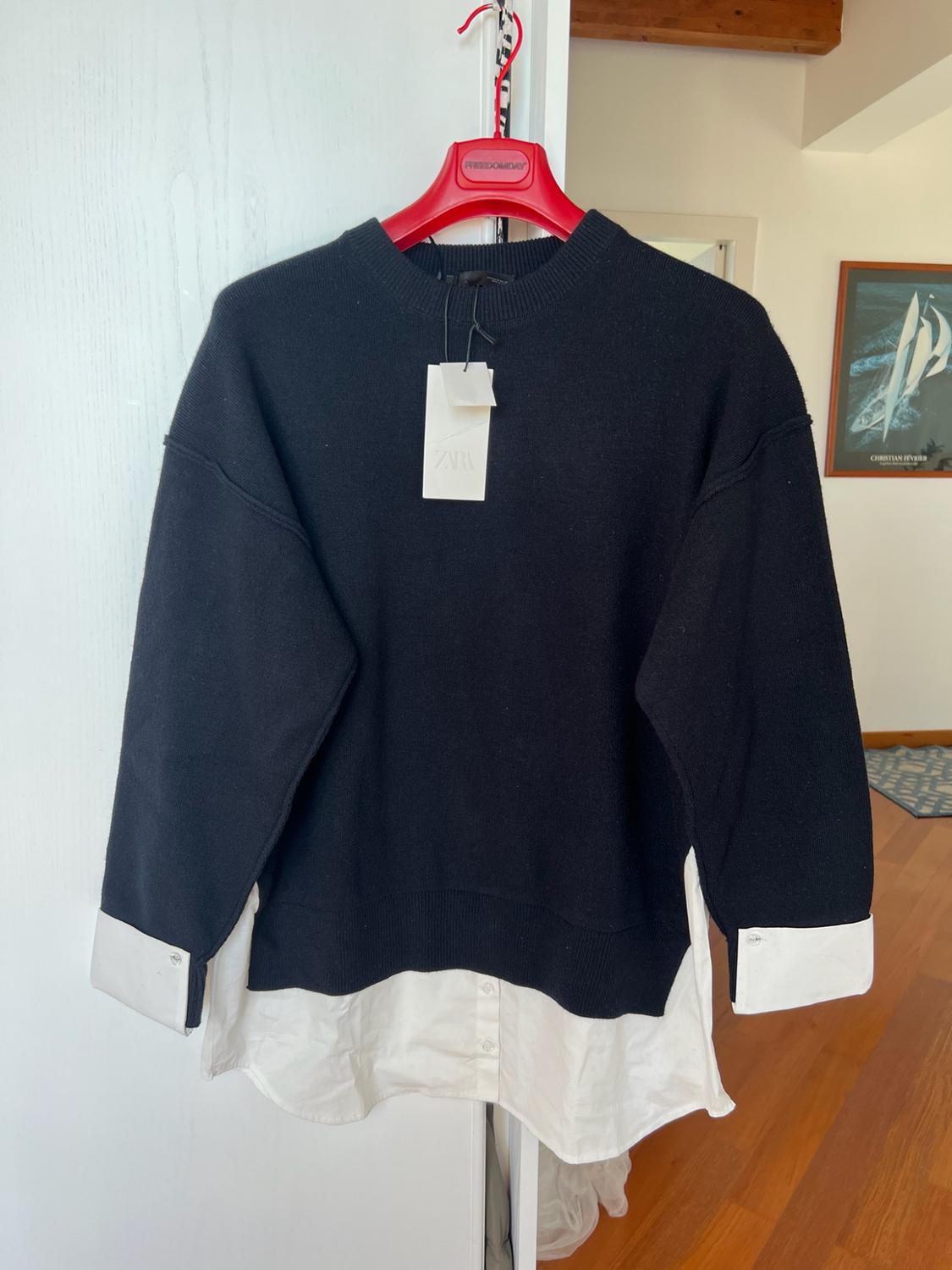 Black jumper with a white shirt hem and cuffs on a red hanger with the text FREEDOMDAY. ZARA label. Room background.