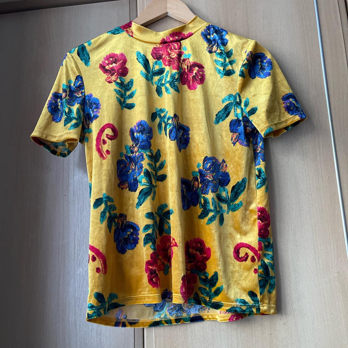 Yellow t-shirt with a floral print in blue, red, and green, hanging on a wooden hanger in front of a light wooden wardrobe.