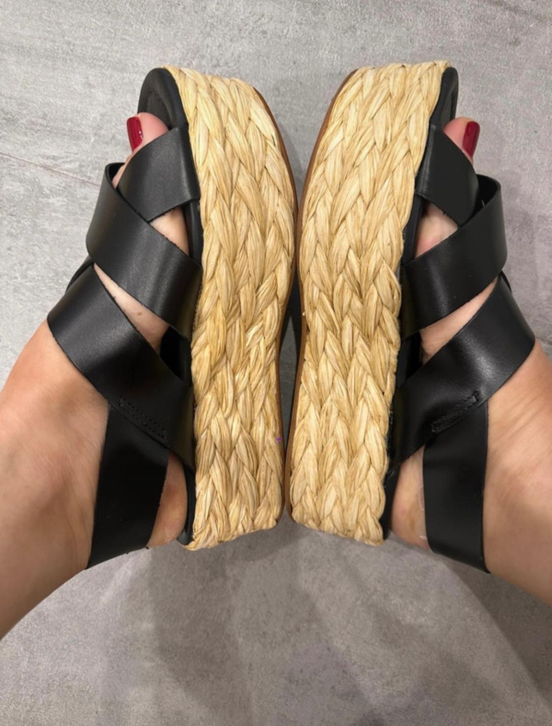 Top-down view of feet with red toenails and black strappy sandals with braided jute platforms on a grey floor.
