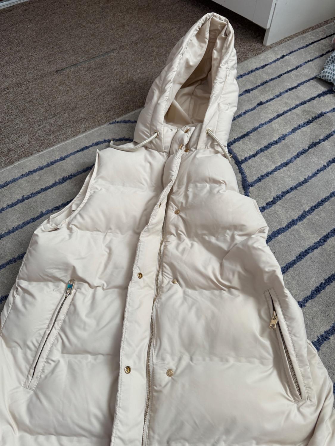 Beige padded gilet with a hood, zip, and pockets, laid out on a light rug with blue stripes in a relaxed setting.