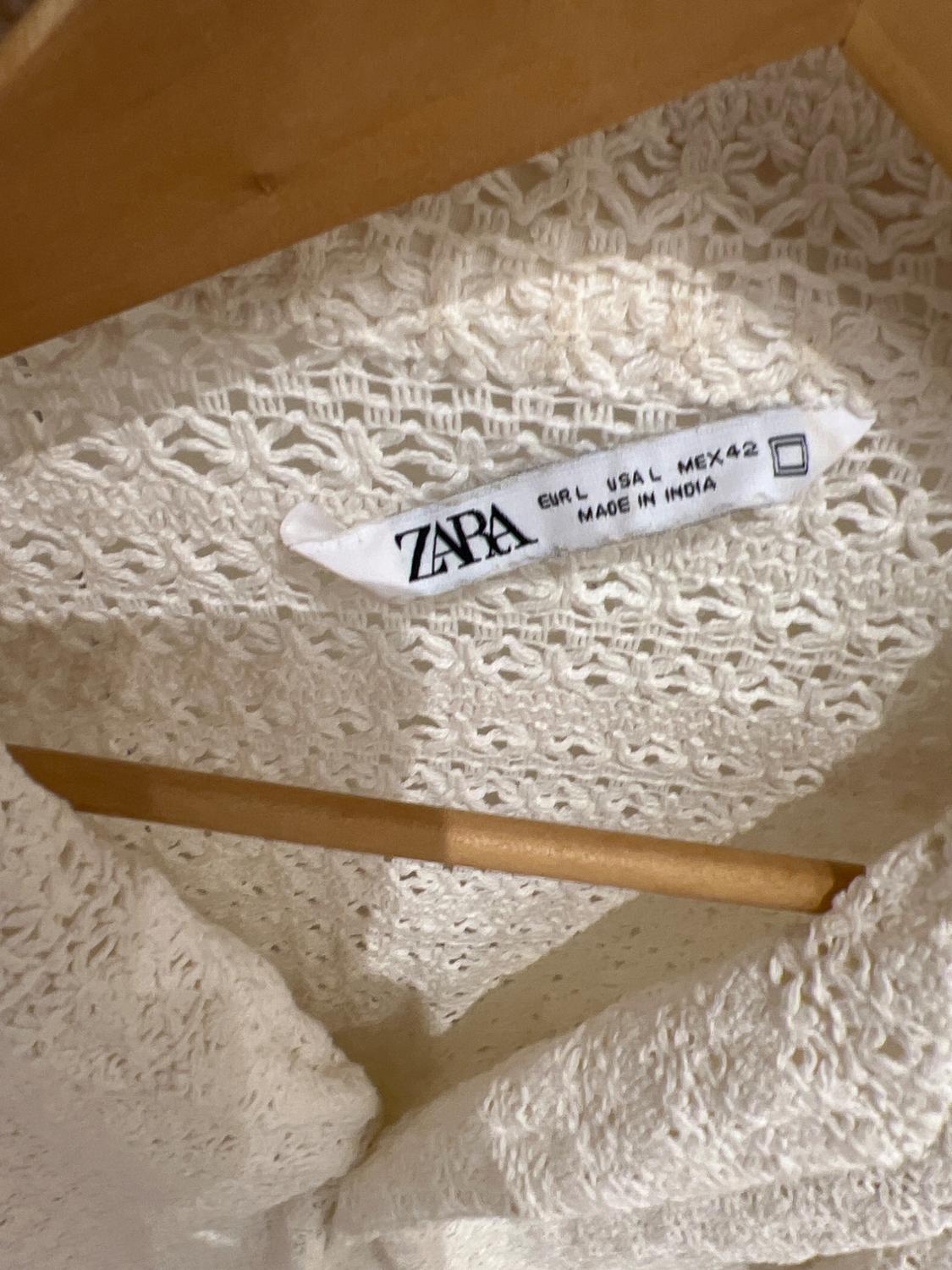White clothing label hanging on a wooden hanger. Visible text: "ZARA EUR L USA L MEX 42 MADE IN INDIA". Fabric with a lace texture.