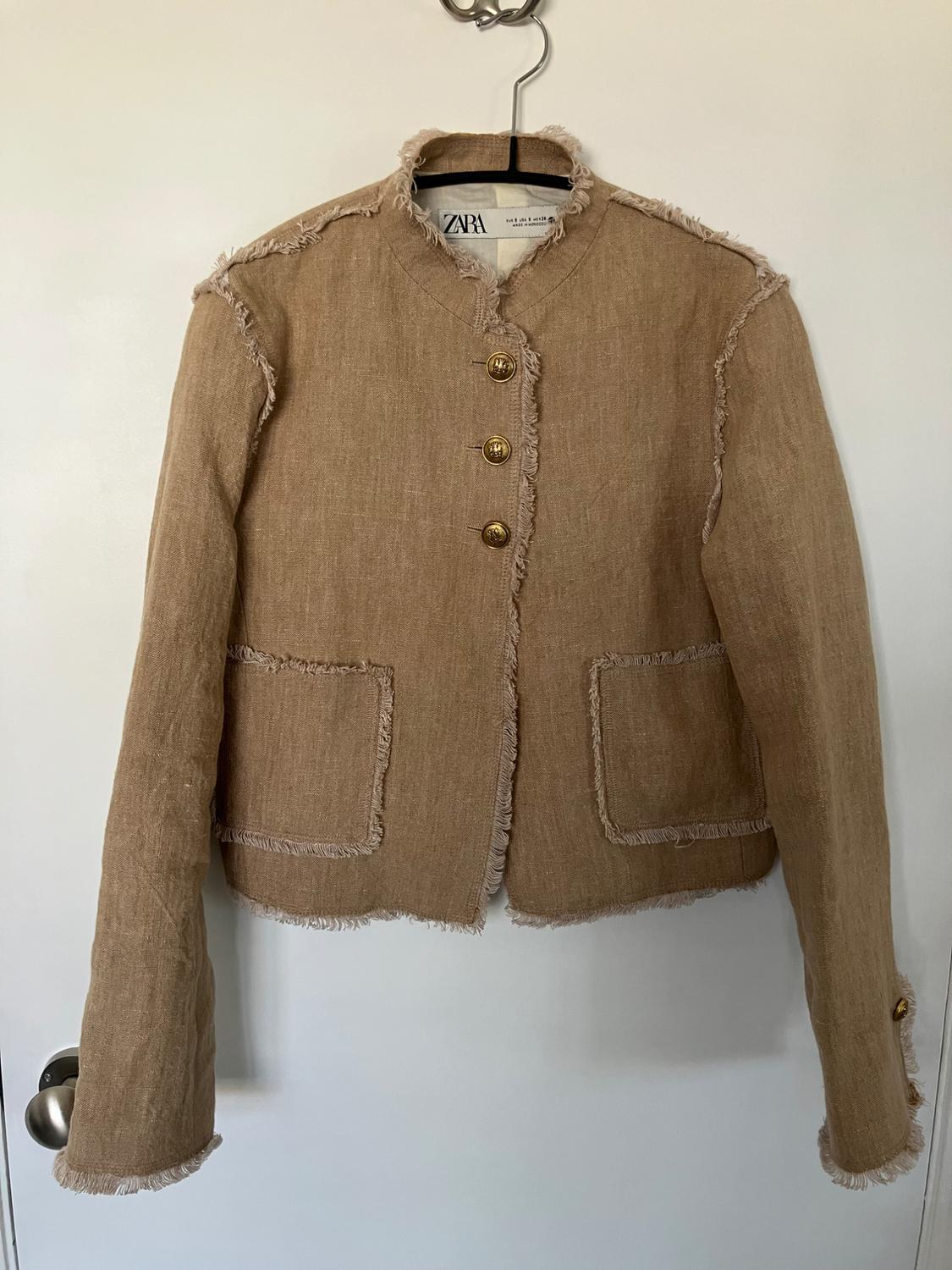 Beige jacket with gold buttons and frayed edges, hanging on a hanger over a white door. Visible tag: ZARA.