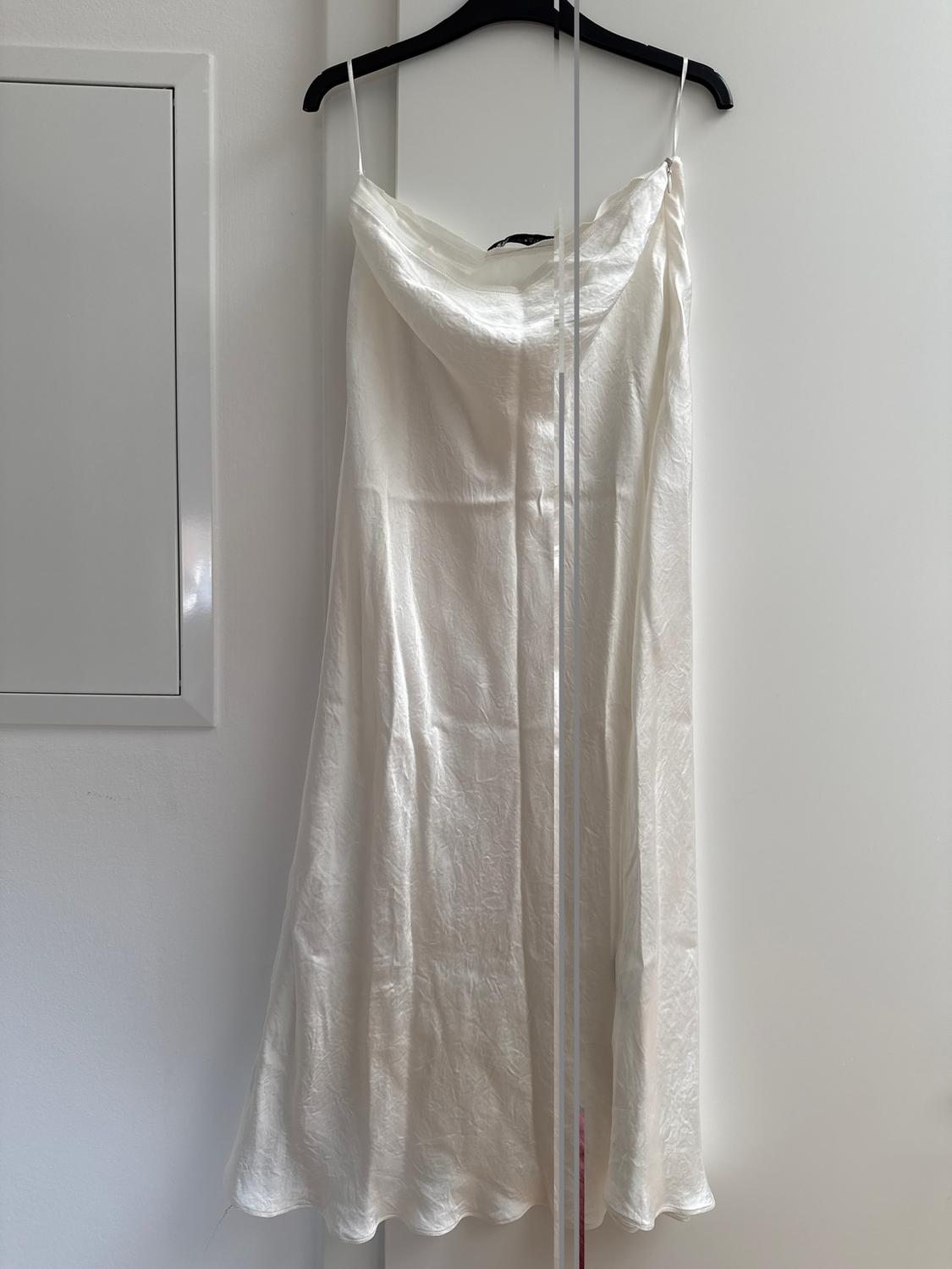 White, thin-strapped dress hanging on a black hanger against a white wall. Reflection in a mirror next to it.