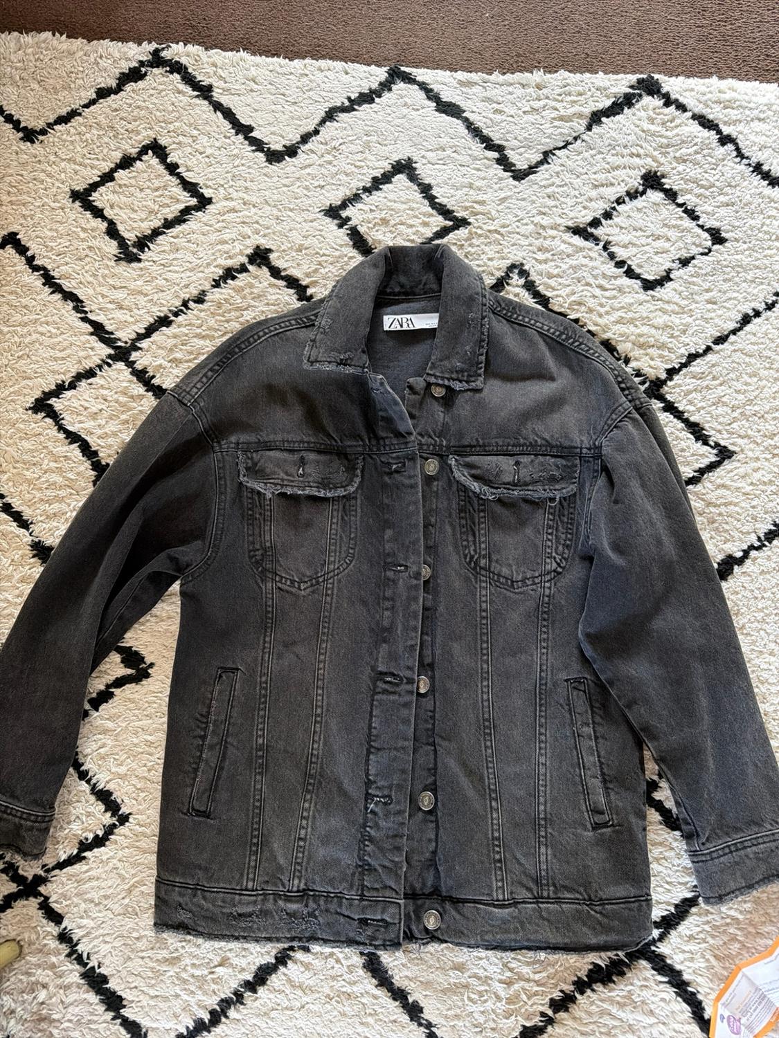 Distressed black denim jacket with a ZARA label on the collar, laid out on a white rug with a black geometric pattern.
