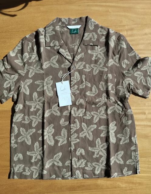 Short-sleeved brown shirt with a beige leaf pattern on a wooden table. White tag hanging from the collar.