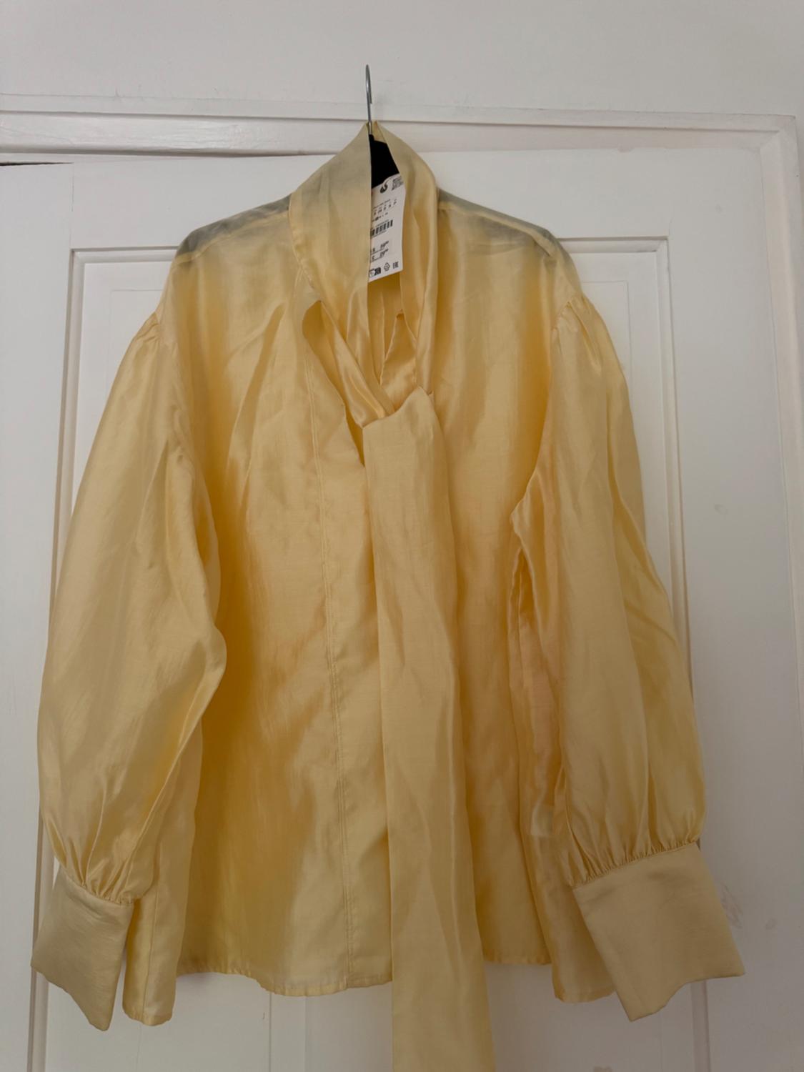 Yellow blouse with a neck tie and puff sleeves, hanging on a hanger on a white door. Relaxed style.