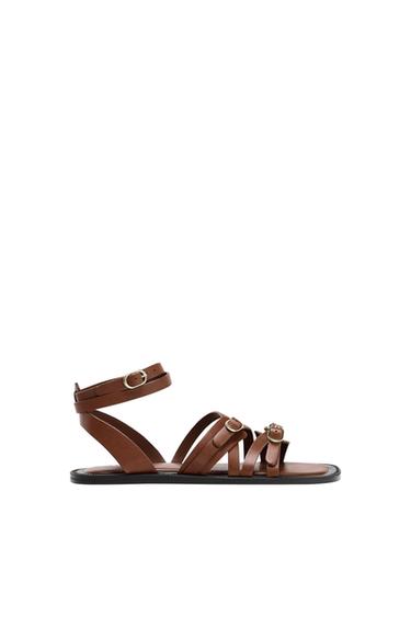 Flat brown strappy sandals with gold buckles on the ankle and front, on a white background.