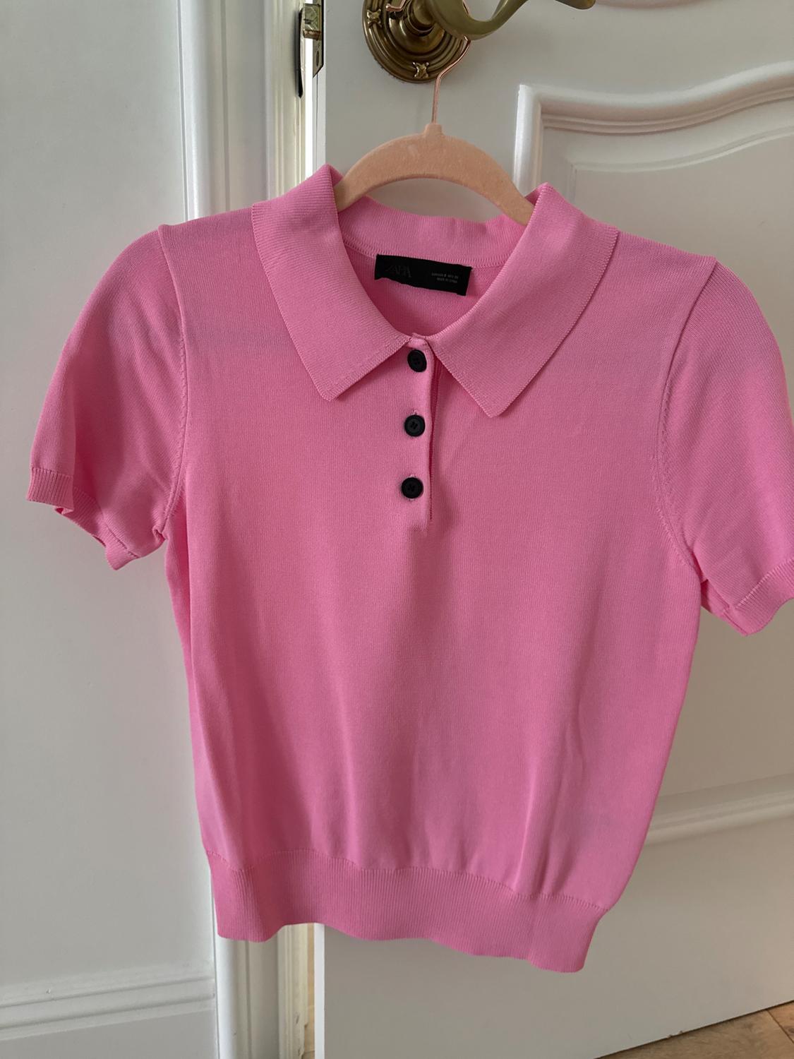 Pink short-sleeved polo shirt with black buttons, hanging on a wooden hanger in front of a white door with a gold handle.